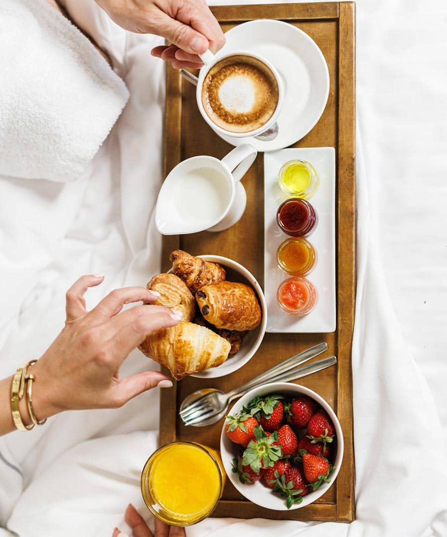 Bandeja con desayuno encima de la cama y dos manos cogiendo un cruasán y un café